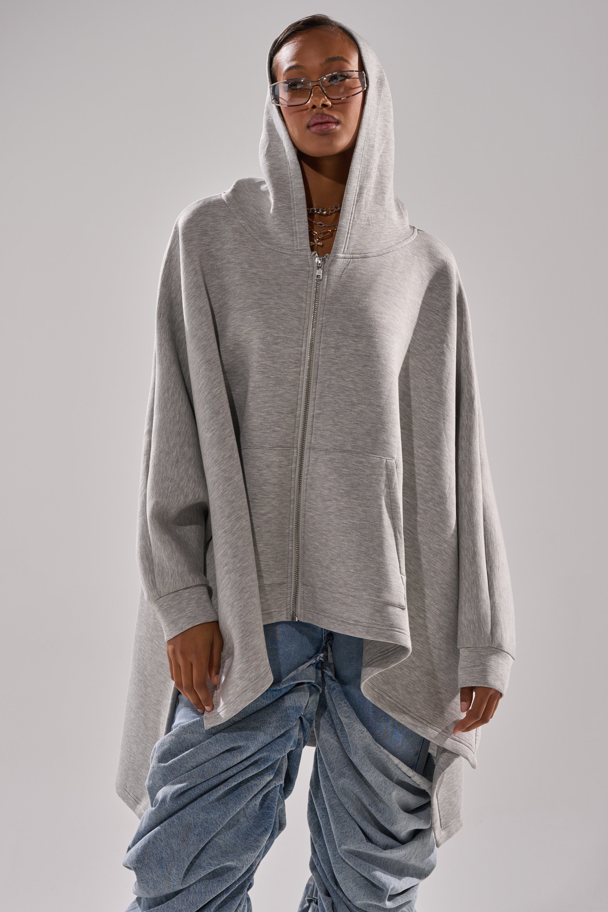 A person wearing the CREW LOVER OVERSIZED ZIP UP SWEATSHIRT in heather grey and ruched blue jeans poses with a neutral expression against a plain gray background.