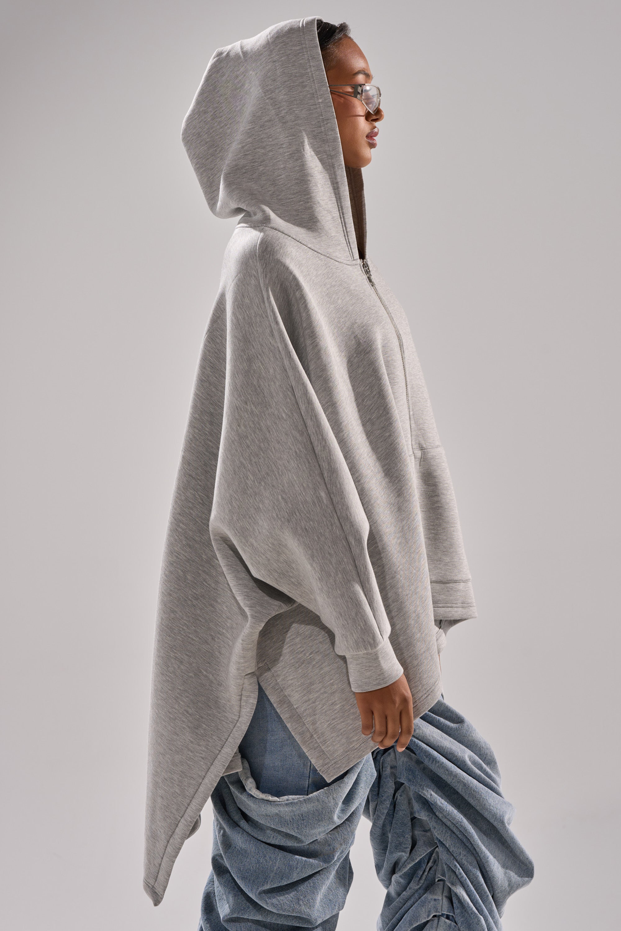 A person in glasses stands in profile against a plain background, wearing a light gray hooded poncho over the CREW LOVER OVERSIZED ZIP UP SWEATSHIRT and ruched blue jeans, with their hands at their sides.