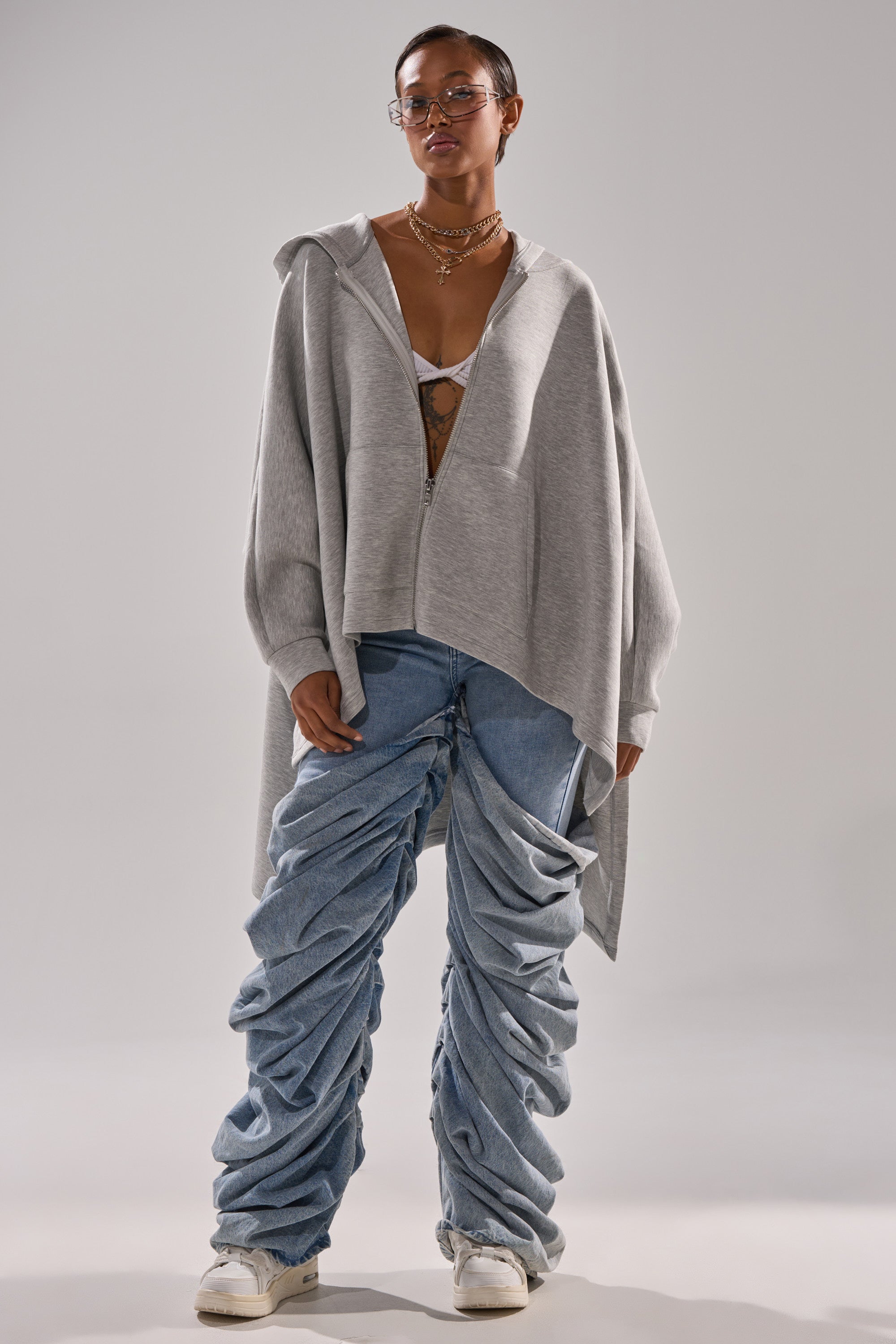 A person wears the CREW LOVER OVERSIZED ZIP UP SWEATSHIRT in heather grey over a white bralette, styled with blue ruched baggy jeans and white sneakers. They accessorize with glasses and layered necklaces against a plain light background.