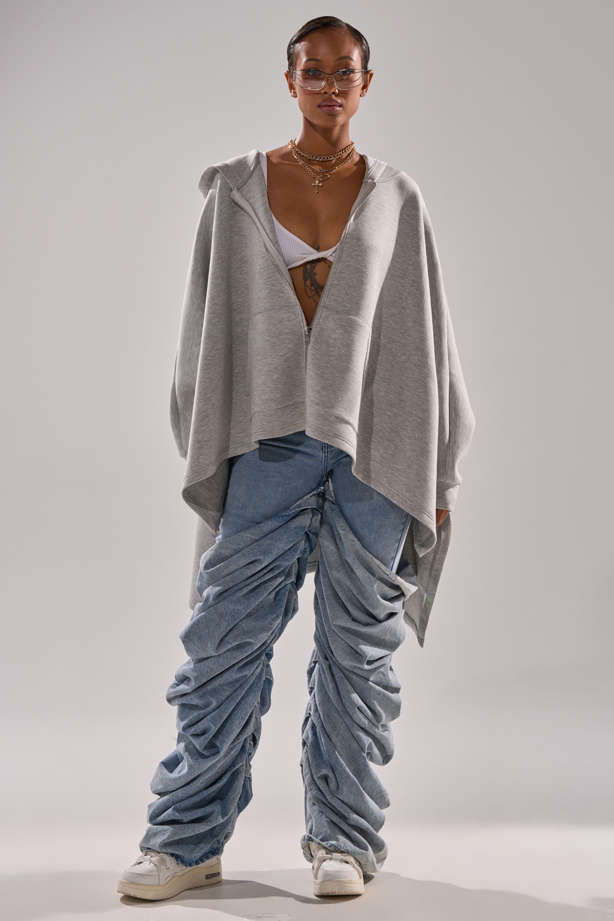 Wearing the CREW LOVER OVERSIZED ZIP UP SWEATSHIRT over a white bralette, layered necklaces, light blue ruched baggy jeans, and white sneakers, this person stands against a plain background.