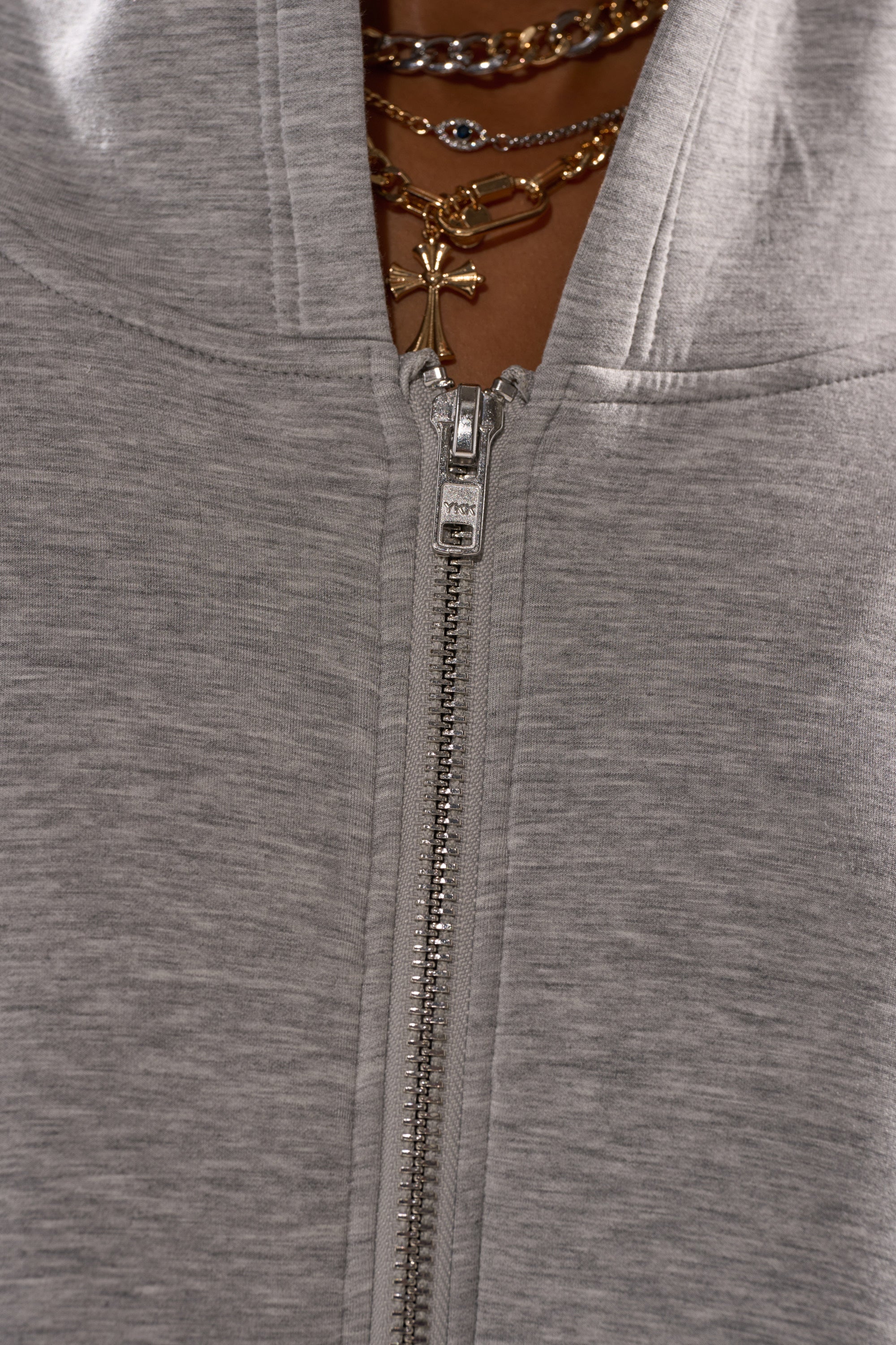 A close-up of the CREW LOVER OVERSIZED ZIP UP SWEATSHIRT in heather grey, partially unzipped to showcase layered gold necklaces with a cross pendant around the neck.