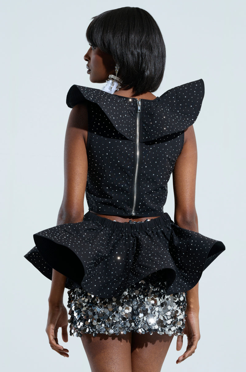 A person wearing the MS FASHIONISTA SLEEVELESS RHINESTONE BLOUSE, accompanied by a sequined skirt and featuring large shoulder accents, showcases their style in a black, textured outfit. The ensemble is completed with bold earrings and shoulder-length hair, all set against a plain background.