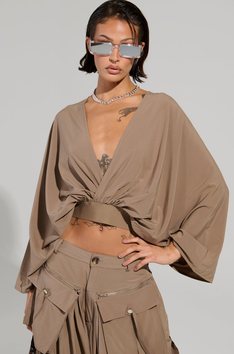 Sporting a pair of reflective sunglasses and a necklace, an individual confidently models the AKIRA Label TALK ABOUT ME BLOUSE, which features a plunging V-neckline. Paired with matching cargo pants, this flowing blouse boasts wide sleeves that subtly reveal a torso tattoo as they place their hands on their hips.