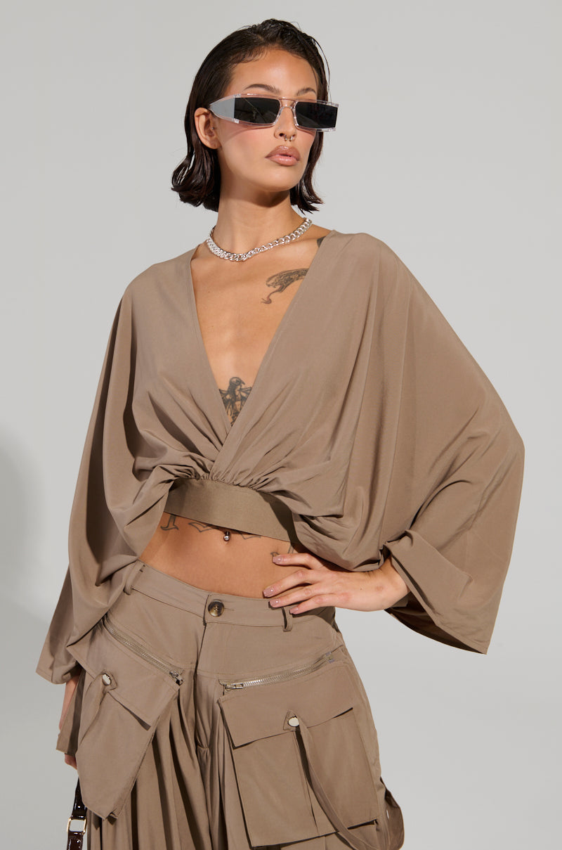 A person with short hair sports rectangular sunglasses and the TALK ABOUT ME BLOUSE by AKIRA Label, which has a plunging V-neckline and wide sleeves. They are paired with beige cargo pants, posing with one hand on their hip, accessorized with a silver necklace and tattoos against a plain background.