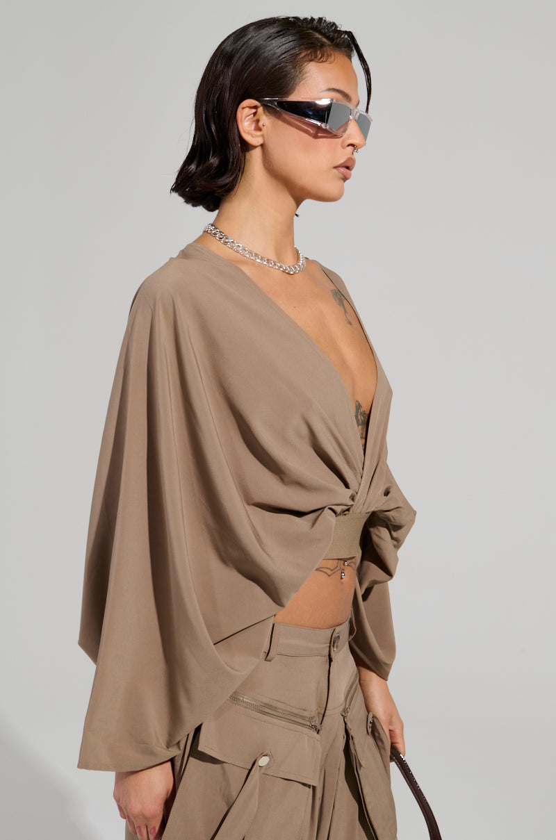 A person wearing angular sunglasses, a silver chain necklace, and the AKIRA Label TALK ABOUT ME BLOUSE with wide sleeves. The taupe top boasts a plunging V-neckline. They have dark, slicked-back hair and carry a light brown bag. Their pants feature multiple zippers and pockets.