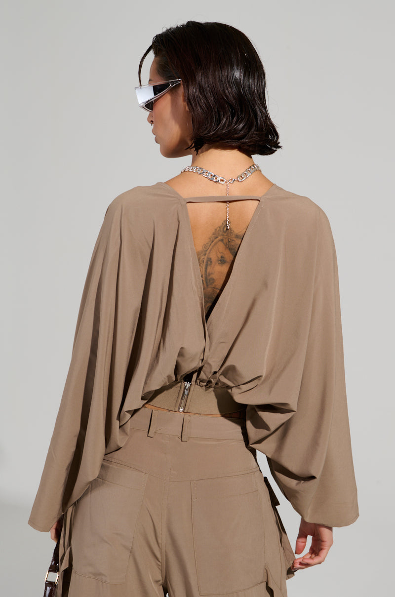 A person with short dark hair is dressed in a light brown ensemble that highlights the TALK ABOUT ME BLOUSE from the AKIRA Label. This draped blouse boasts a plunging v-neckline and an open back, showcasing a tattoo. They accessorize their stylish look with sunglasses and a chain necklace while posing against a plain backdrop.