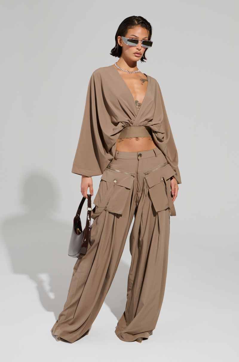 Dressed in taupe-toned pants with oversized pockets and the AKIRA Label TALK ABOUT ME BLOUSE, which has wide sleeves and a plunging v-neckline, this person accessorizes with sunglasses and holds a brown bag, all set against a simple, light-colored backdrop.