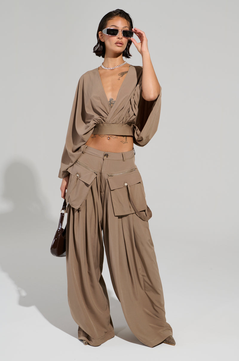 Dressed in a fashionable tan outfit with wide-legged pants and the TALK ABOUT ME BLOUSE from AKIRA Label, an individual stands confidently. The blouse features a wrap style with a plunging V-neckline and semi-stretchy knit fabric, adding to their sophisticated appearance. Complemented by sunglasses, a small handbag, and short hair, they radiate contemporary elegance.