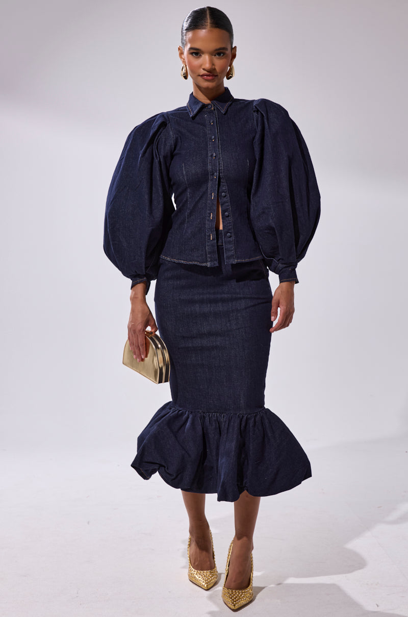 A woman stands in a studio wearing the LET'S CHANGE IT UP DENIM MIDI SKIRT with a button-up top featuring puffed sleeves. She accessorizes with a gold clutch, gold heels, and large hoop earrings.