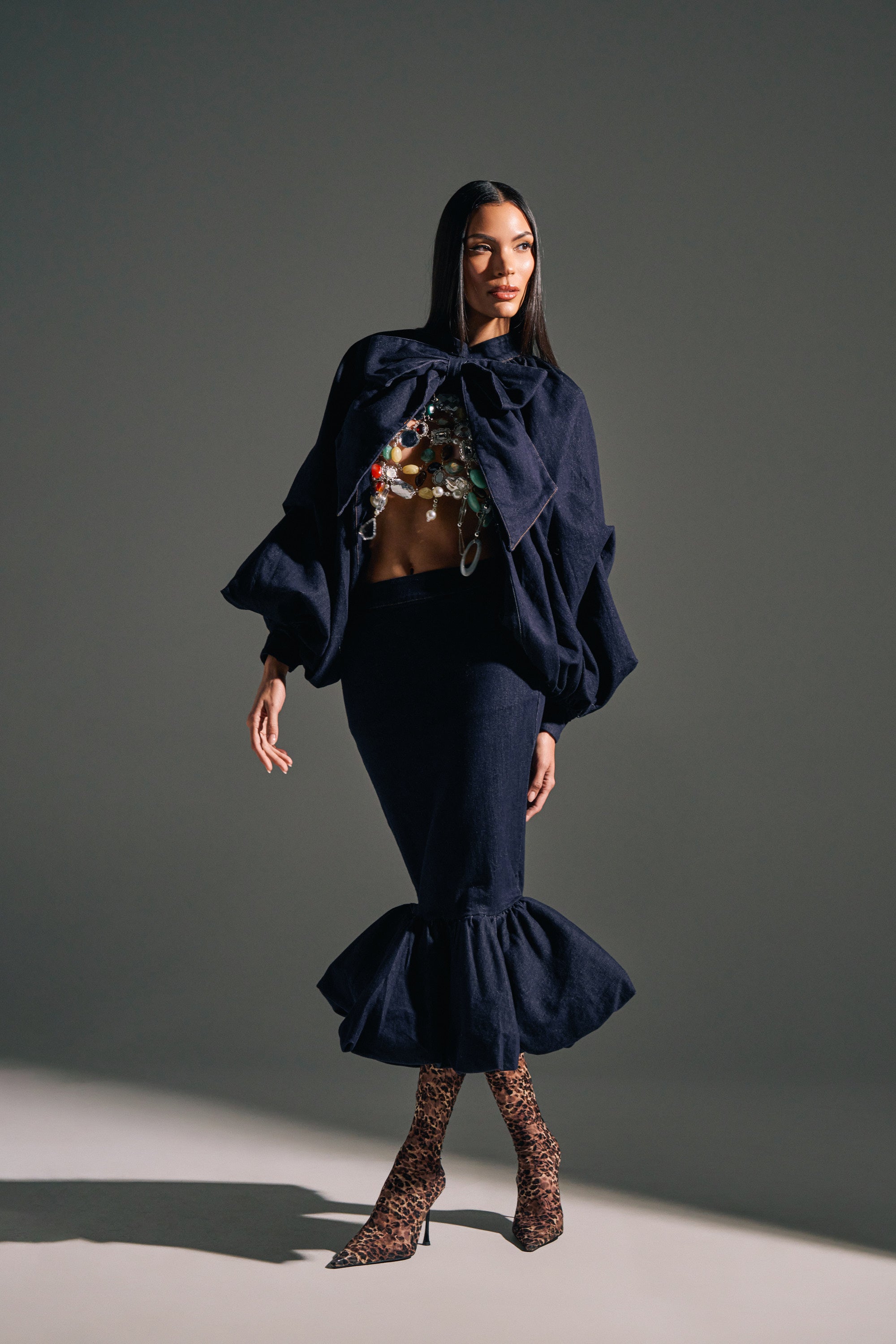 A woman poses against a neutral background wearing the LET'S CHANGE IT UP DENIM MIDI SKIRT, styled with a cropped cape, embellished top, and animal print heeled boots.