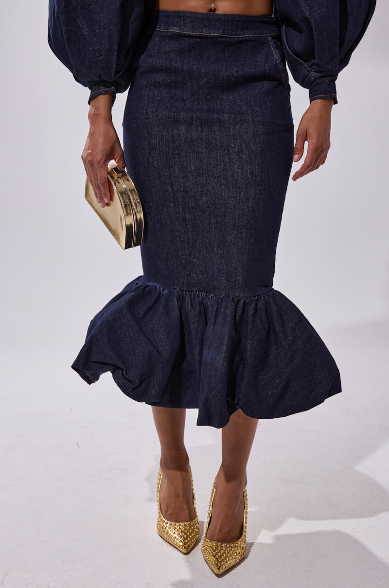 Wearing the LET'S CHANGE IT UP DENIM MIDI SKIRT with a flounced, ruffled hem, puffed-sleeve top, gold studded heels, and a gold clutch, a person stands against a plain light background.