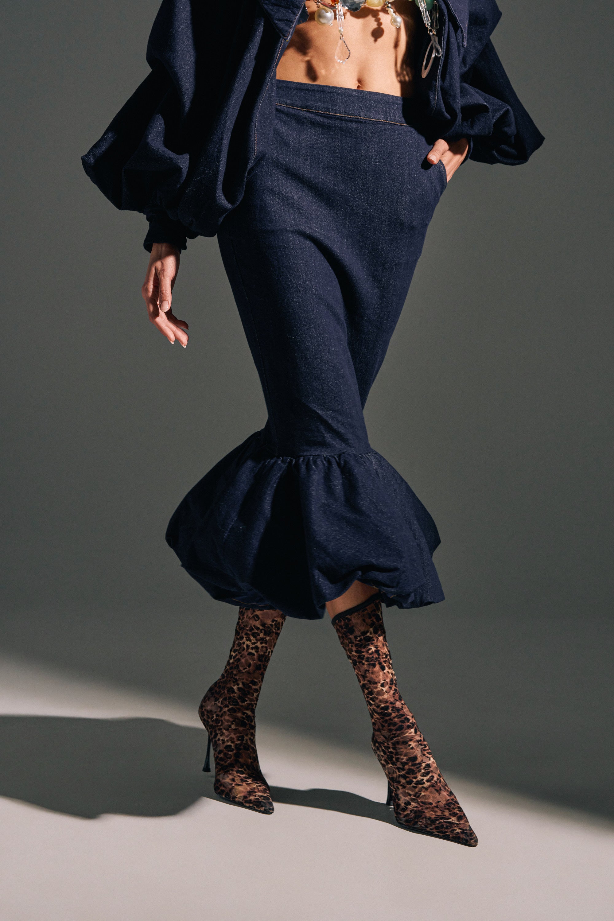 Wearing the LET'S CHANGE IT UP DENIM MIDI SKIRT with a cropped top and sheer leopard-print heeled boots, a person poses against a plain background with one hand in their pocket.