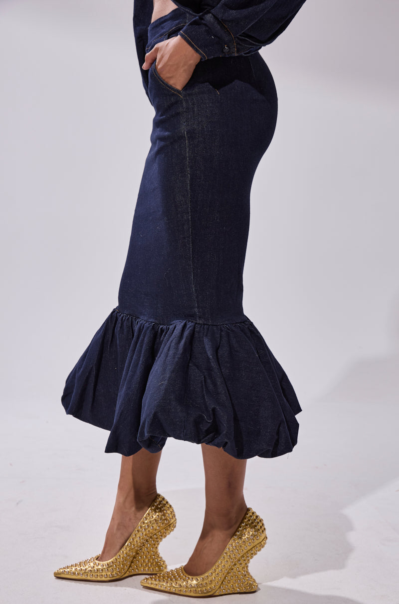 Person wearing the LET'S CHANGE IT UP DENIM MIDI SKIRT with a ruffled hem, paired with gold textured high heels and a matching denim top, hand in pocket, against a plain light background.