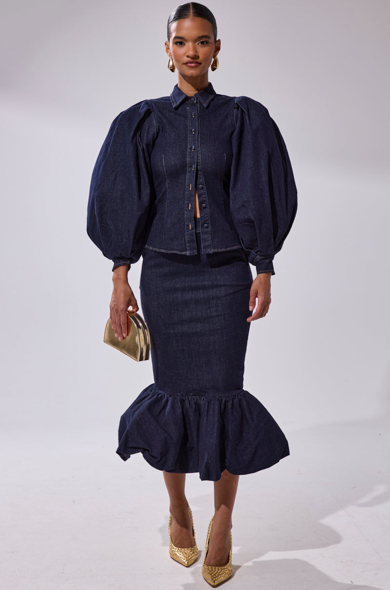 A woman models the LET'S CHANGE IT UP DENIM MIDI SKIRT, a high-waisted dark denim skirt with a ruffled hem, paired with a puff-sleeve button-up top. She accessorizes with gold heels and a matching clutch against a plain background.