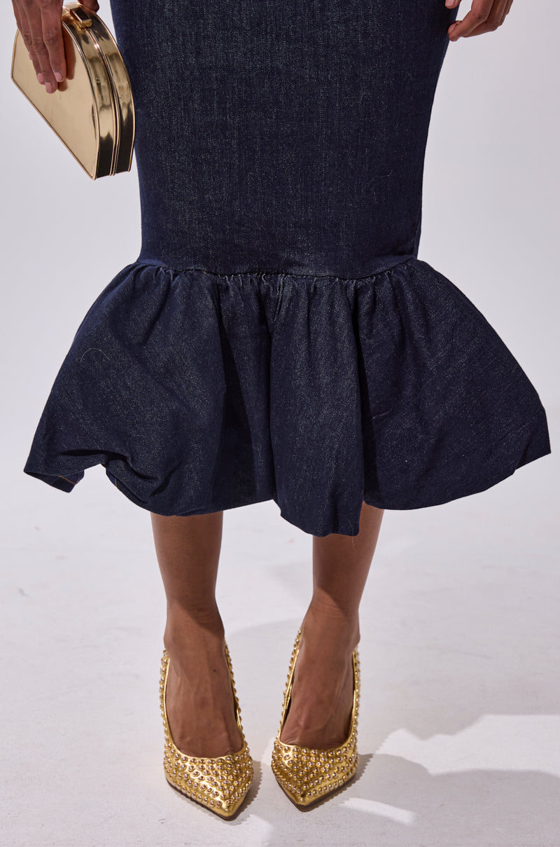 A person wearing the LET'S CHANGE IT UP DENIM MIDI SKIRT with a ruffled hem, gold studded high heels, and holding a shiny gold clutch stands against a plain light-colored background.