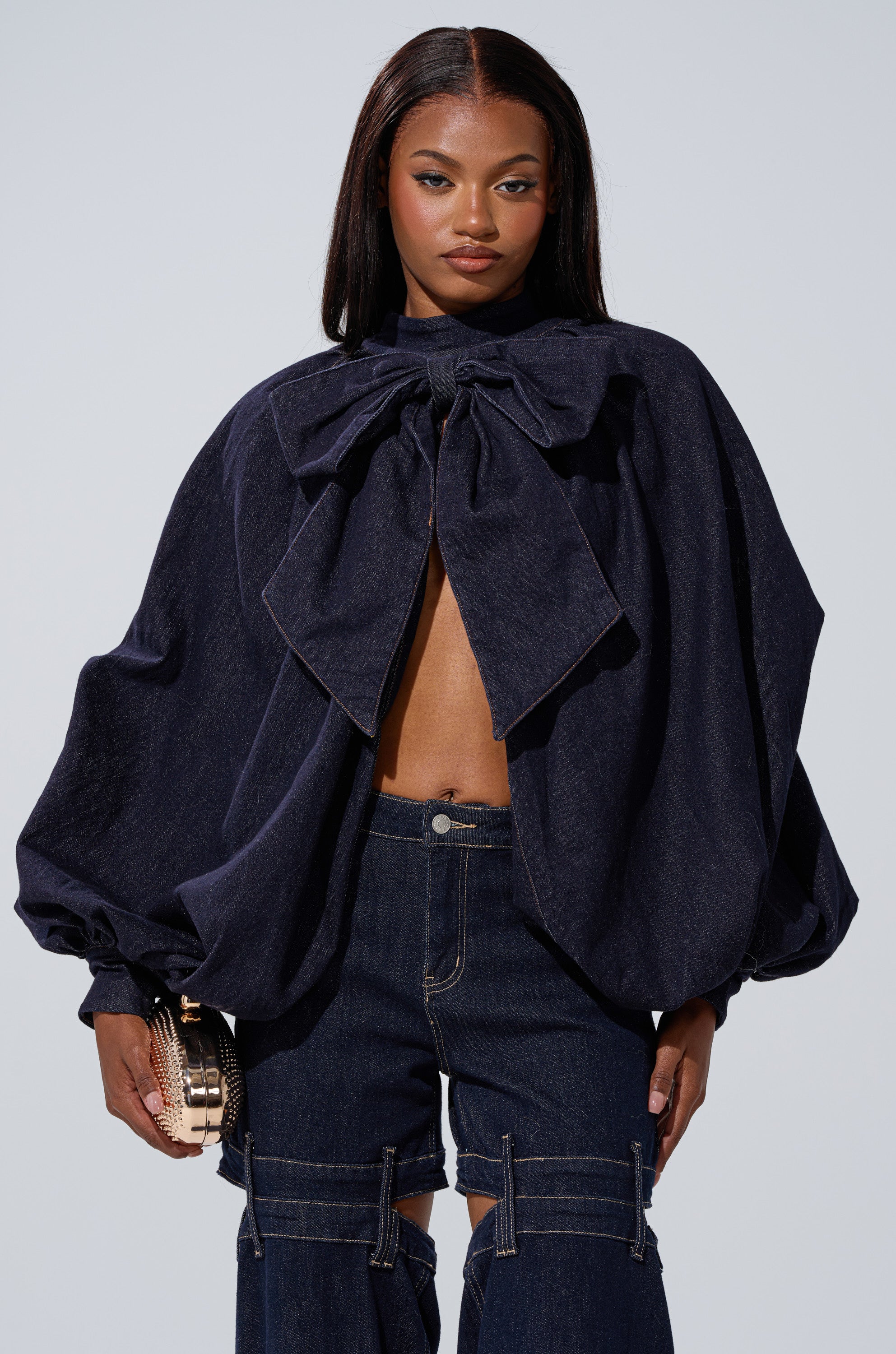 A woman in wide-legged dark denim pants and the LEAH Ruched Denim Mock Neck Top in dark blue, featuring voluminous sleeves and a large bow, holds a small gold purse against a plain light background.