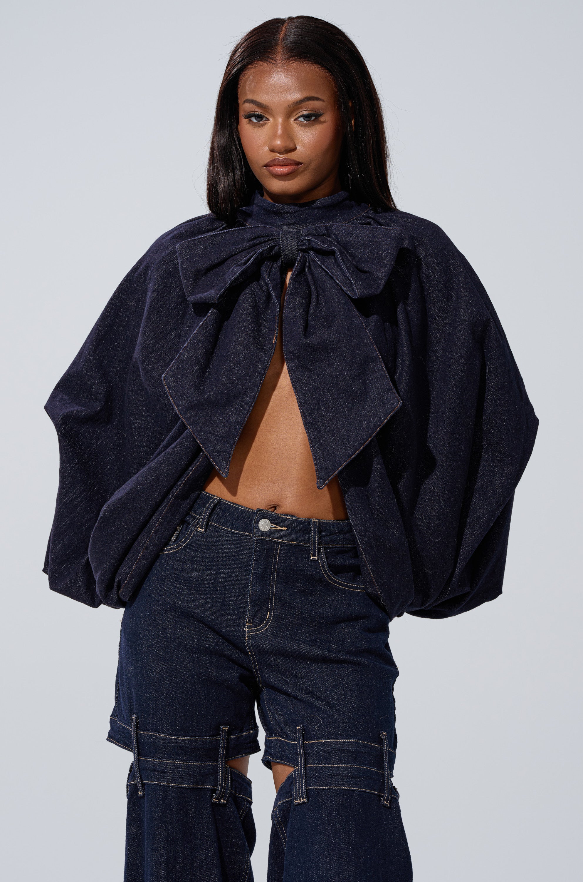 A woman models the LEAH Ruched Denim Mock Neck Top in dark blue denim, styled with voluminous sleeves and a large bow, paired with matching wide-leg pants featuring visible seams and knee cut-outs.