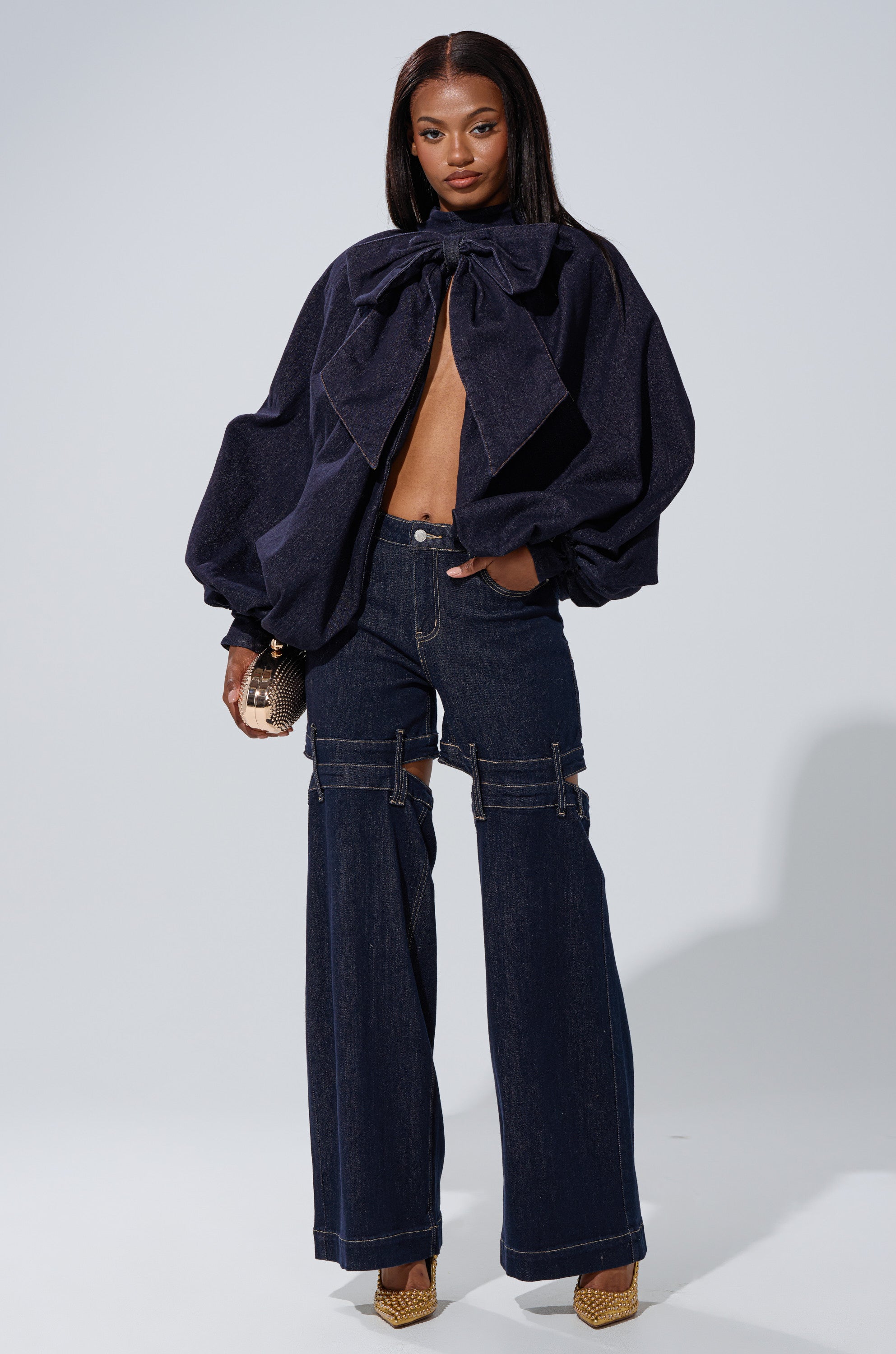 A woman stands confidently in the LEAH Ruched Denim Mock Neck Top in Dark Blue Denim, wide-leg dark blue jeans with zipper details, gold heels, and a small textured clutch against a plain light background.