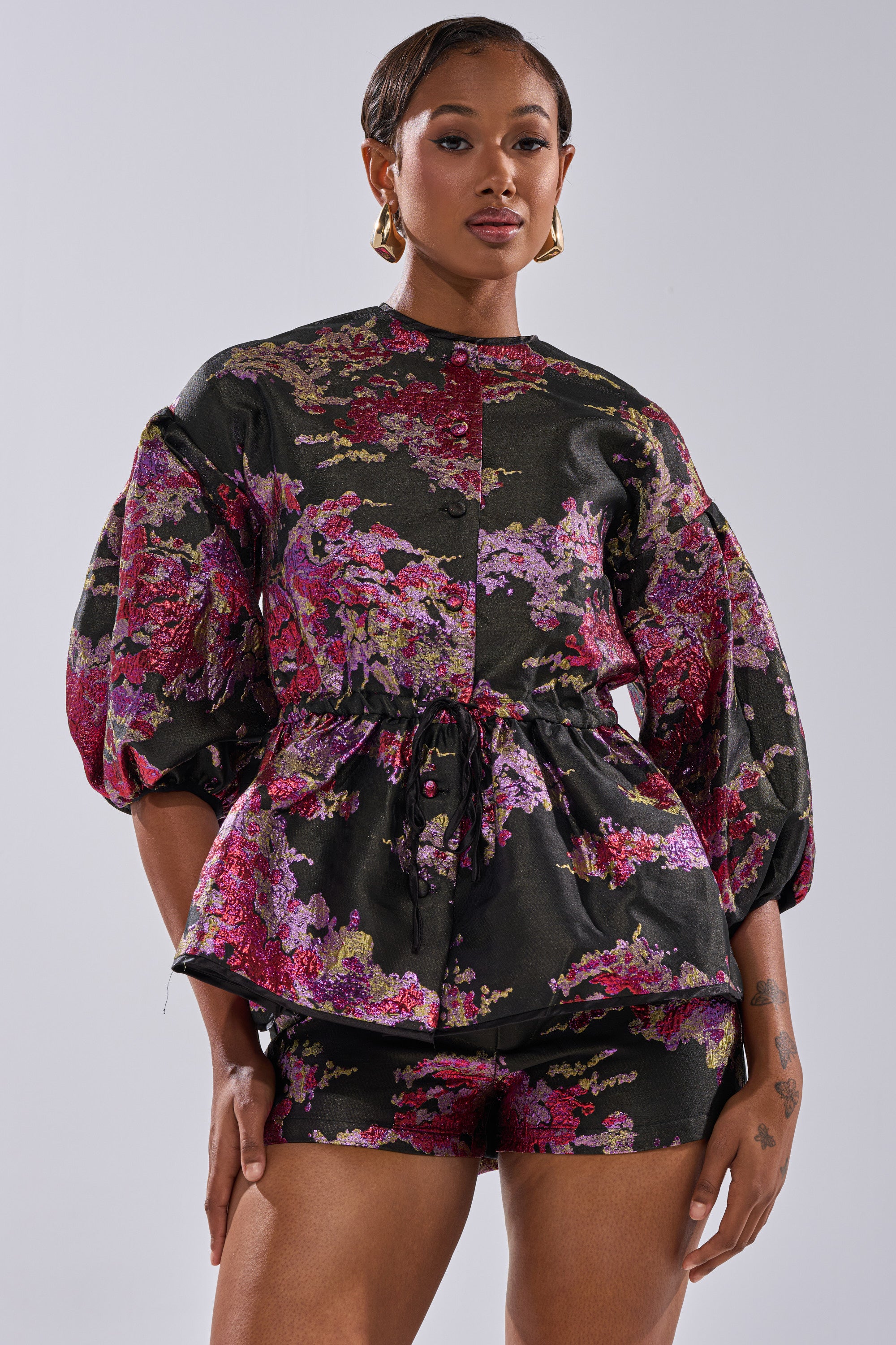 A woman poses confidently in the ETHEREAL BROCADE SHORT SLEEVE BLOUSE IN PINK, styled with matching shorts. The vintage-inspired look features puffed sleeves, a cinched waist, and bold floral patterns. Gold hoop earrings and short hair complete her chic style.