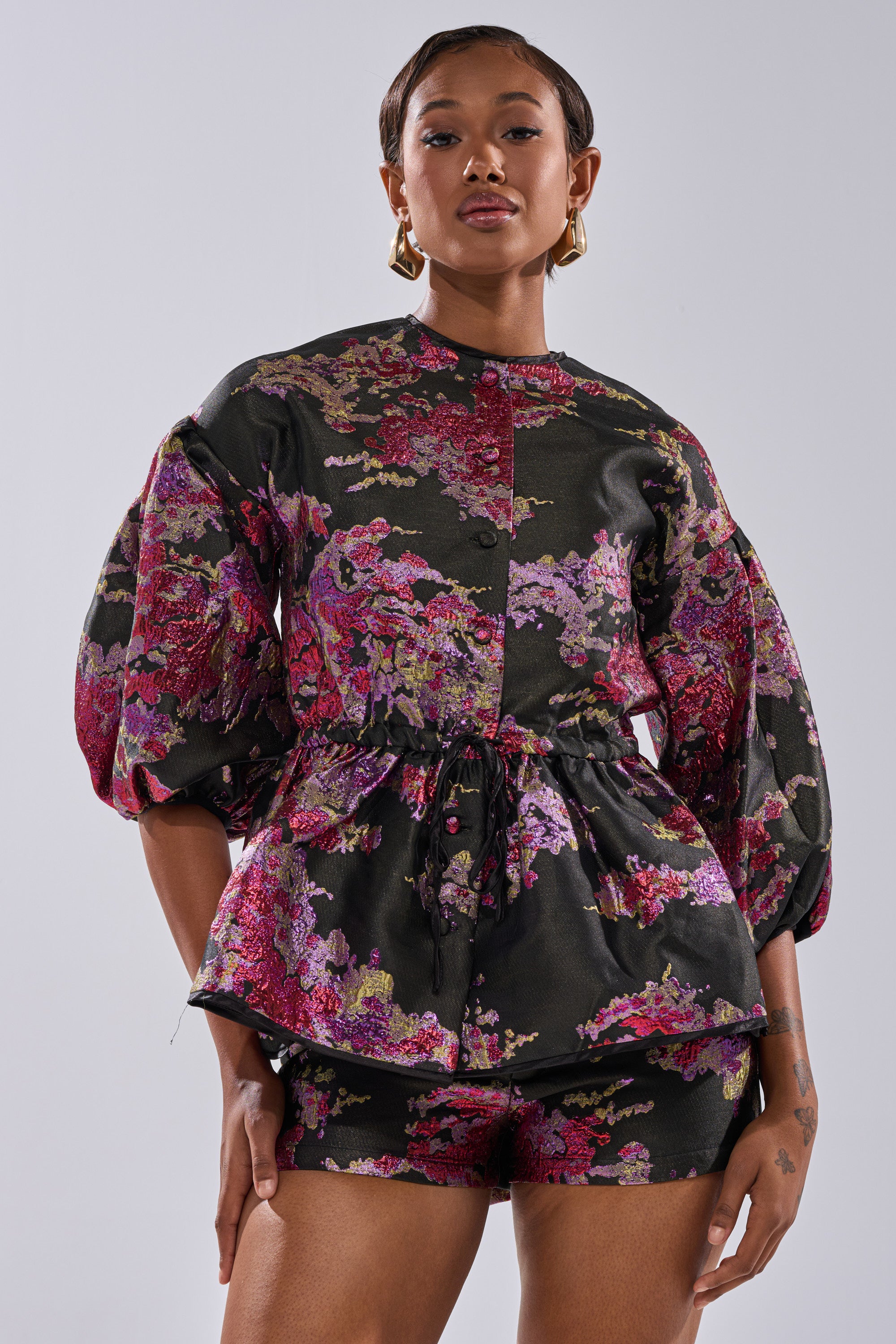 A woman poses confidently against a plain light gray background, wearing the ETHEREAL BROCADE SHORT SLEEVE BLOUSE IN PINK with matching shorts.