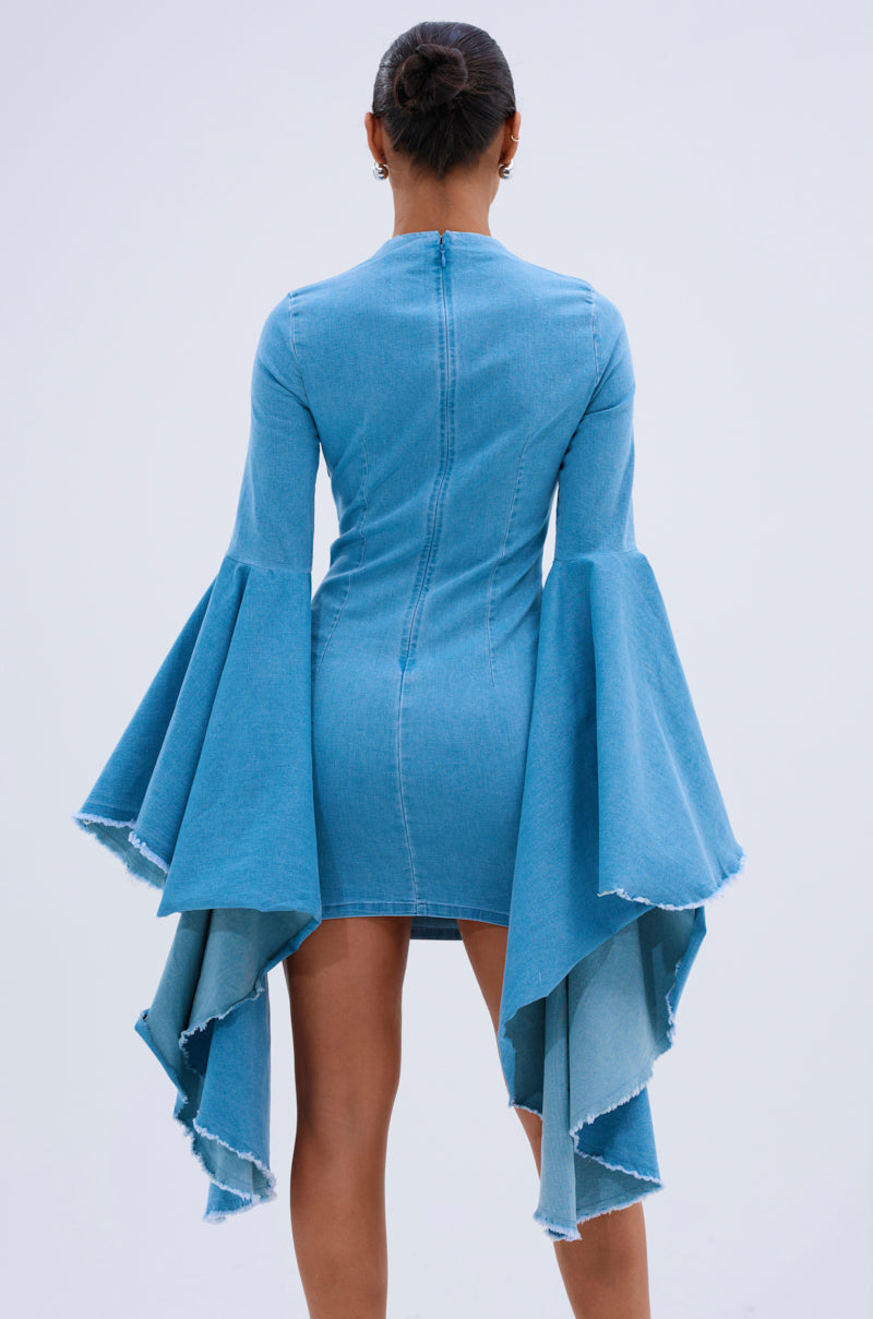 A woman stands facing away, wearing the SHE’S ROYAL DENIM LONG SLEEVE MINI DRESS. The bodycon mini features dramatic bell sleeves and asymmetrical wing-like skirt panels, flattering her silhouette as her hair is styled in a neat bun.