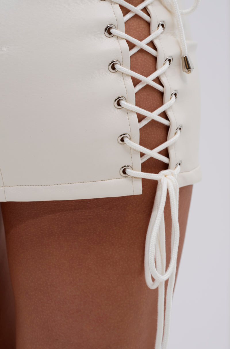 Close-up of a person wearing the FESTIVAL GIRLY LACE UP MINI SKIRT in white faux leather with lace-up sides, white cord through metal eyelets, and skin peeking beneath the lacing—perfect for on-trend festival style.