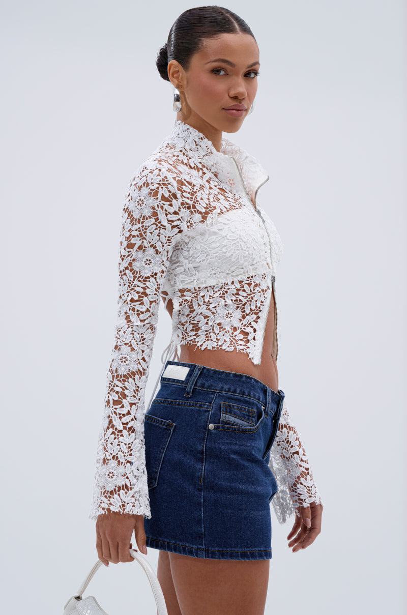 A woman wears the GIMME MORE LACE ZIP-UP TOP in white with long sleeves, paired with a dark denim mini skirt and holding a small white bag, posing sideways against a plain light background.