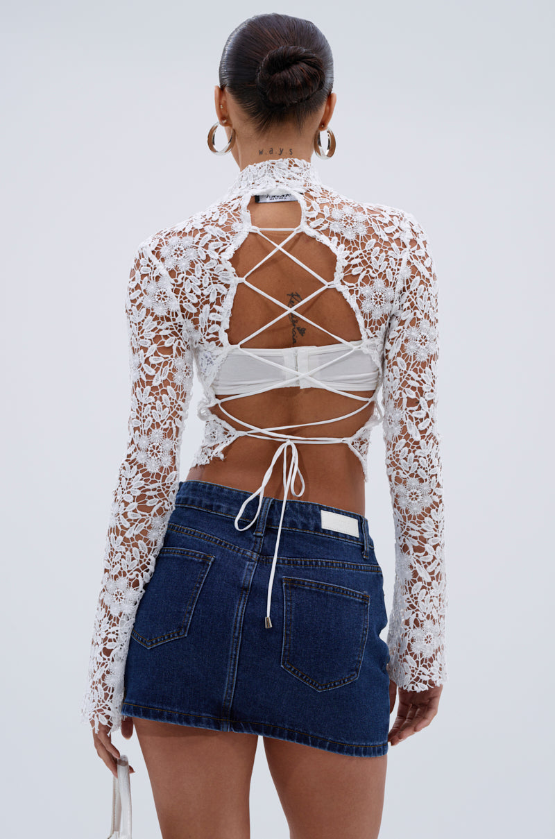 Wearing the GIMME MORE LACE ZIP-UP TOP, featuring white long sleeves and crisscross tie details on the back, paired with a blue denim mini skirt, a person with a sleek bun stands against a plain light background.