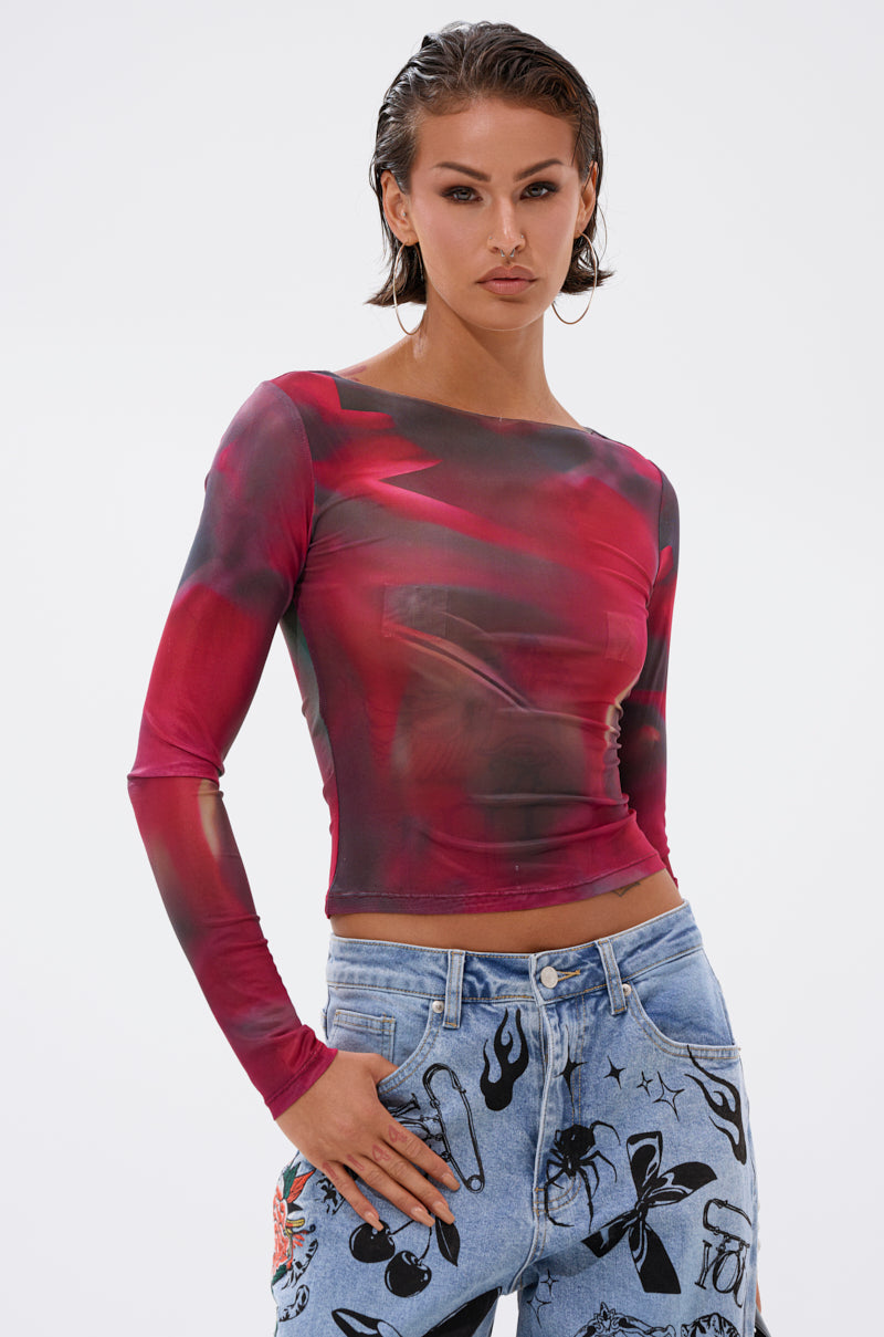 A woman with short, slicked-back hair wears the bold AURA LONG SLEEVE MESH TOP in red and black and light blue jeans with abstract patterns. She stands confidently against a plain white background, looking at the camera.