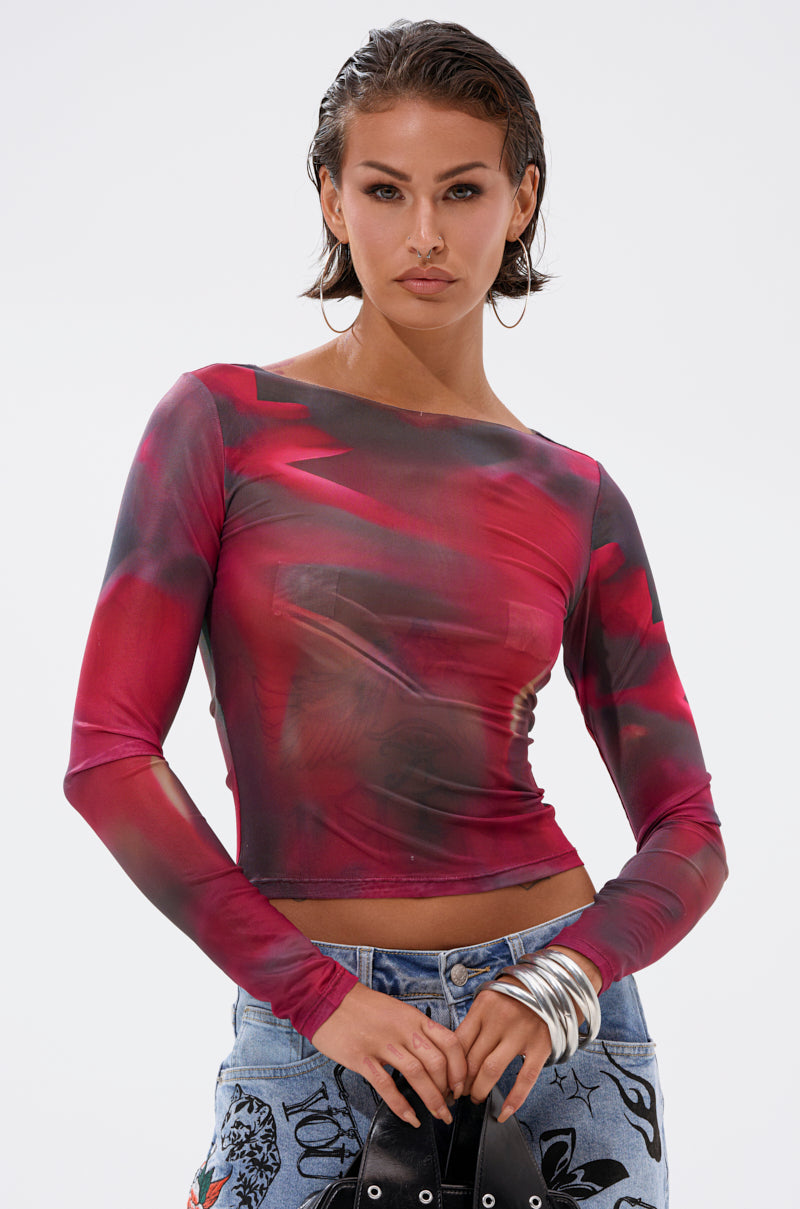 A woman with slicked-back hair models the AURA LONG SLEEVE MESH TOP, featuring sheer mesh and an abstract red and black pattern, paired with light blue embroidered jeans, posing confidently against a plain background.