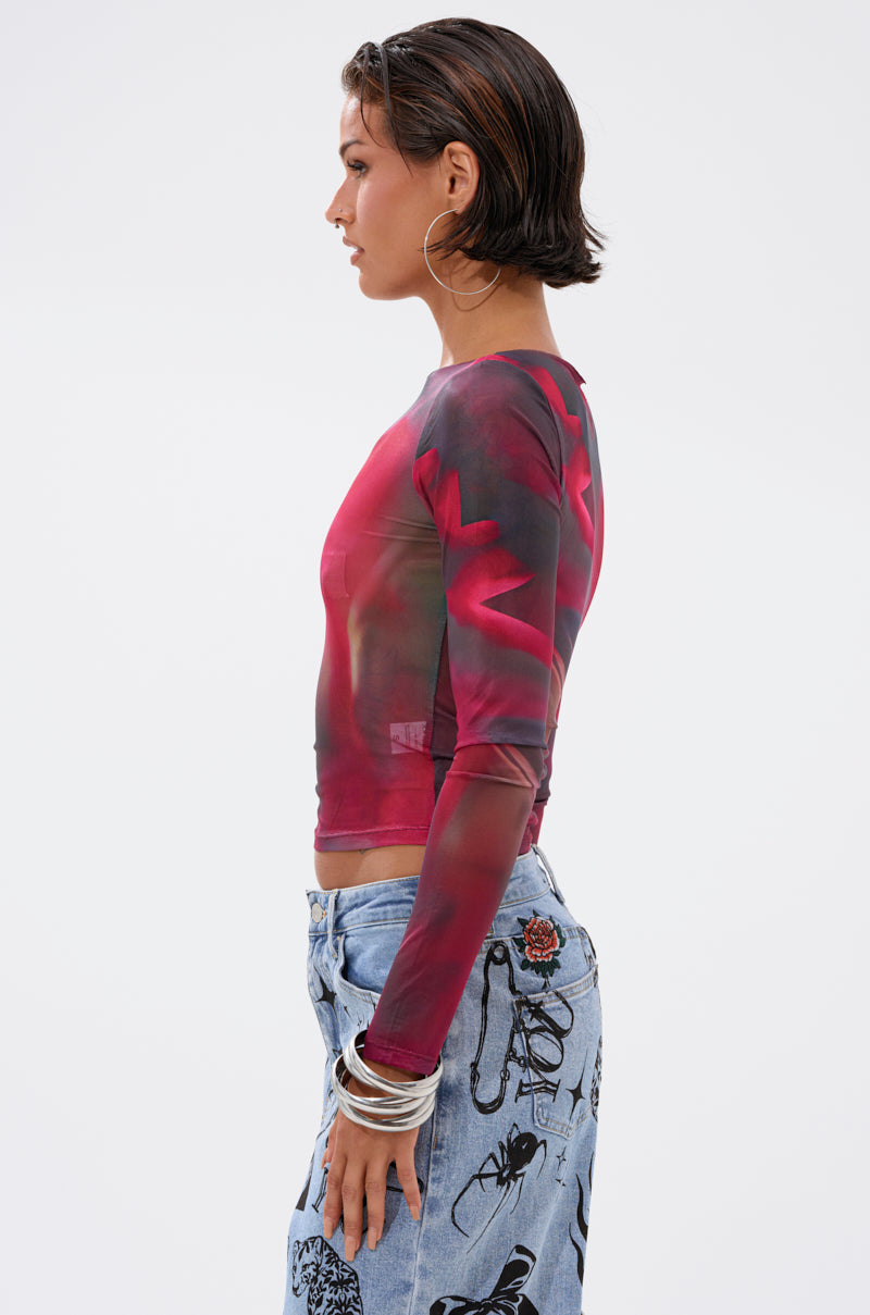 A woman with short brown hair, hoop earrings, and silver bangles wears the AURA LONG SLEEVE MESH TOP—red and black with an abstract pattern—and light blue jeans with black graphic prints, standing in profile against a plain white background.