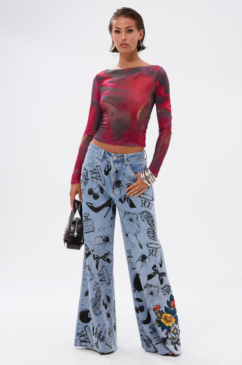 A woman stands against a plain background wearing the AURA LONG SLEEVE MESH TOP and high-waisted, wide-leg blue jeans with black abstract patterns and a colorful floral patch, holding a small black handbag.