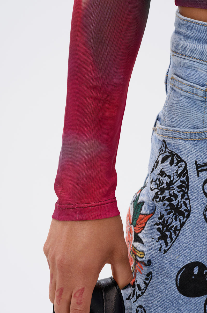 A person wears the AURA LONG SLEEVE MESH TOP in sheer red, paired with light blue jeans embroidered with a cat and leaves, holding a black object against a plain background.