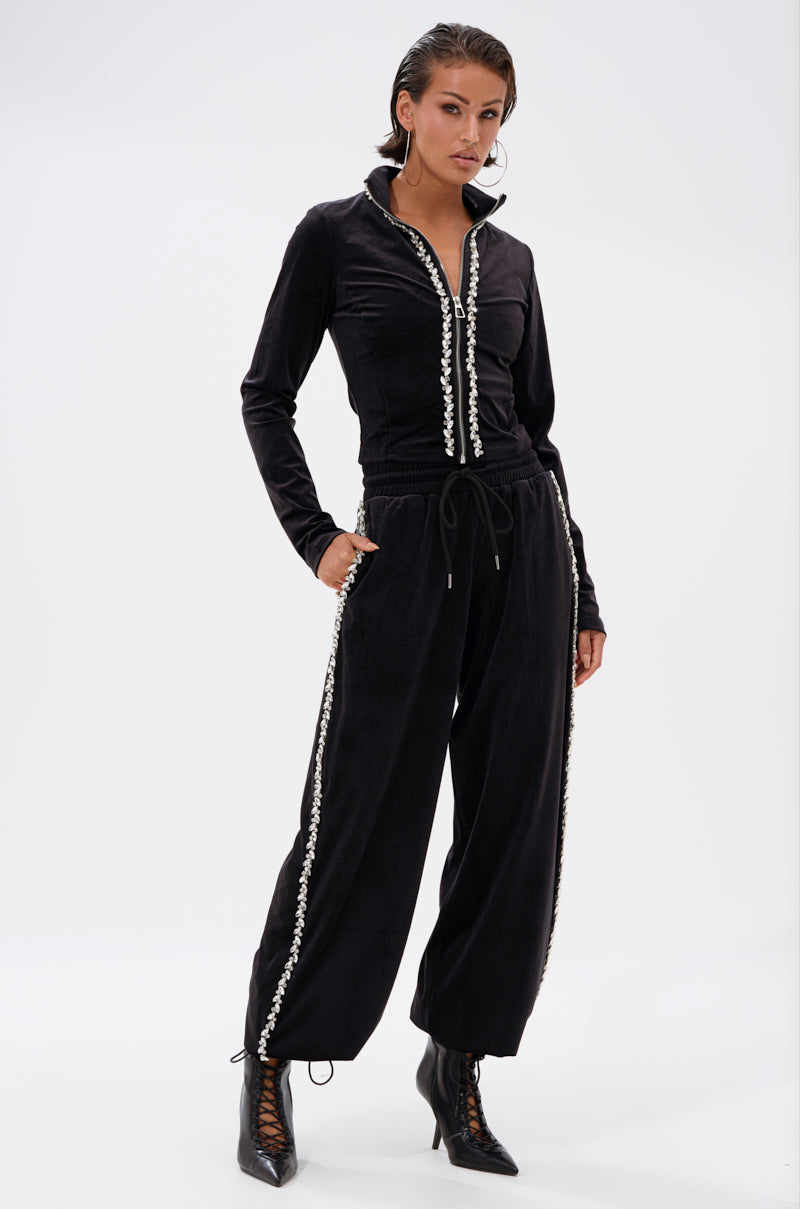 A woman models the DARA RHINESTONE WIDE LEG SWEATPANT, featuring black velour fabric with white trim and rhinestone details. She stands with hands in pockets, wearing black lace-up ankle boots against a plain white background.