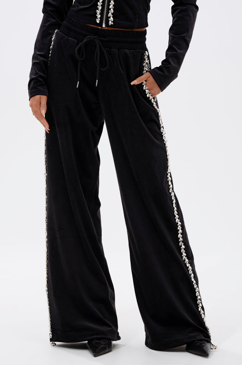 A person wears the DARA RHINESTONE WIDE LEG SWEATPANT—loose-fitting black sweatpants with rhinestone trim on the side seams, an elastic drawstring waistband, and paired with a matching top.