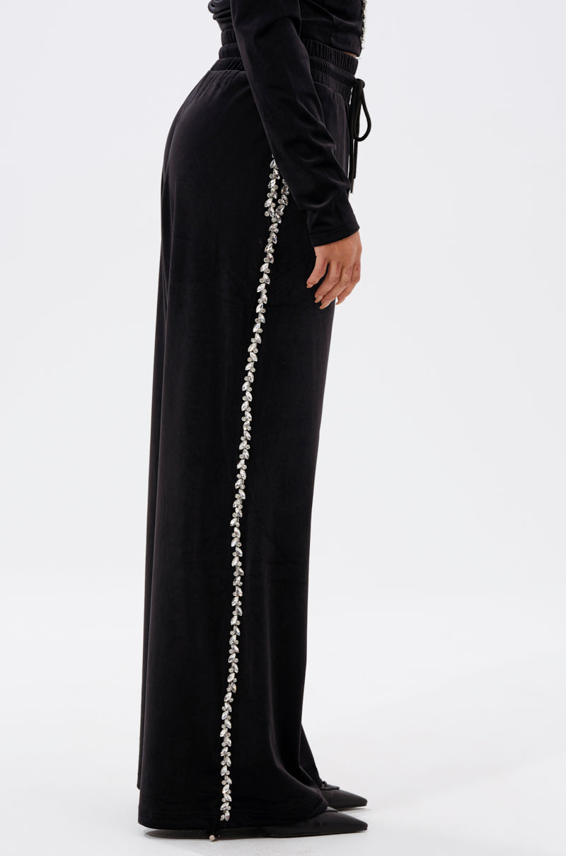A person in DARA RHINESTONE WIDE LEG SWEATPANT, featuring black wide legs and decorative white rhinestone trim along the side seam, stands in a side profile against a plain light background.