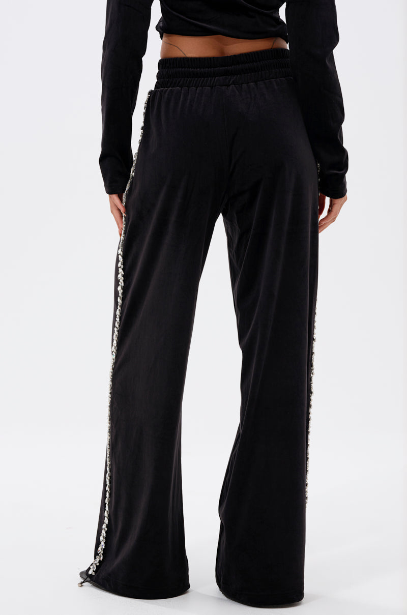 A person wearing the DARA RHINESTONE WIDE LEG SWEATPANT stands facing away, hands at their sides. The black sweatpants have a decorative side stripe, elastic waistband, and zip details at the bottom. Background is plain white.