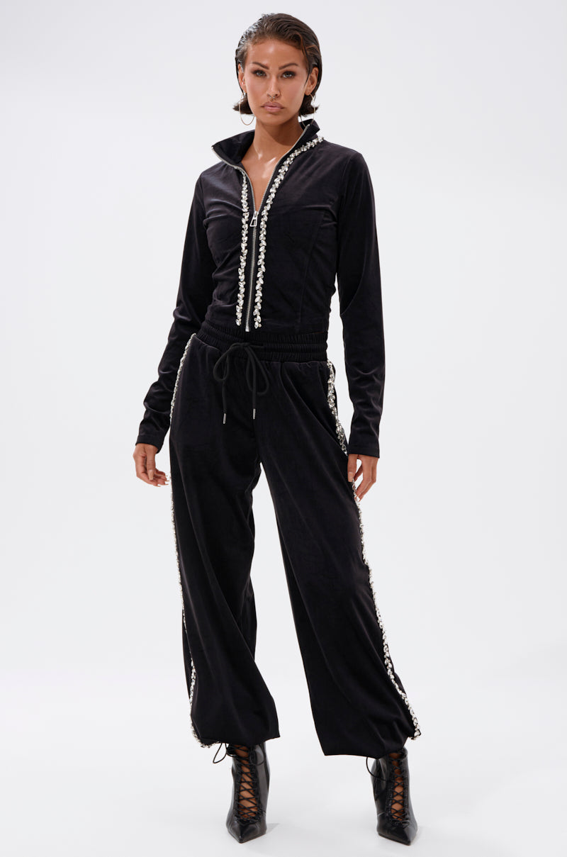 A woman stands confidently in the DARA RHINESTONE WIDE LEG SWEATPANT, featuring white trim along the zipper and pant sides, paired with black lace-up heeled boots against a plain light background.