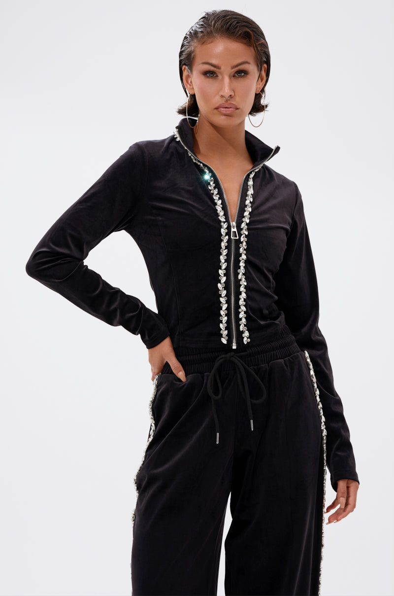 A woman confidently poses with one hand in her pocket, wearing the DARA RHINESTONE MOCK NECK ZIP-UP SWEATSHIRT and matching velour tracksuit pants against a plain background.