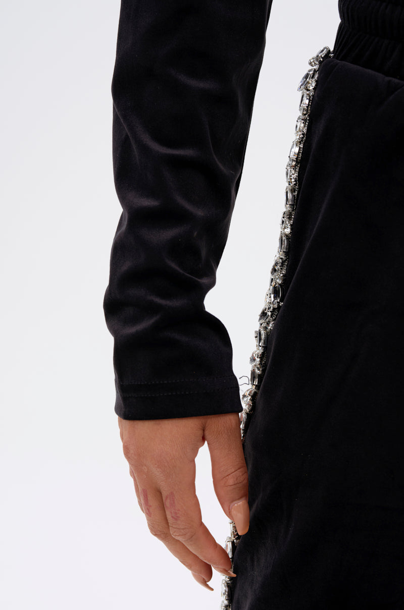A close-up of a person's hand resting by their side, wearing the DARA RHINESTONE MOCK NECK ZIP-UP SWEATSHIRT in black velour with jeweled trim against a plain light background.