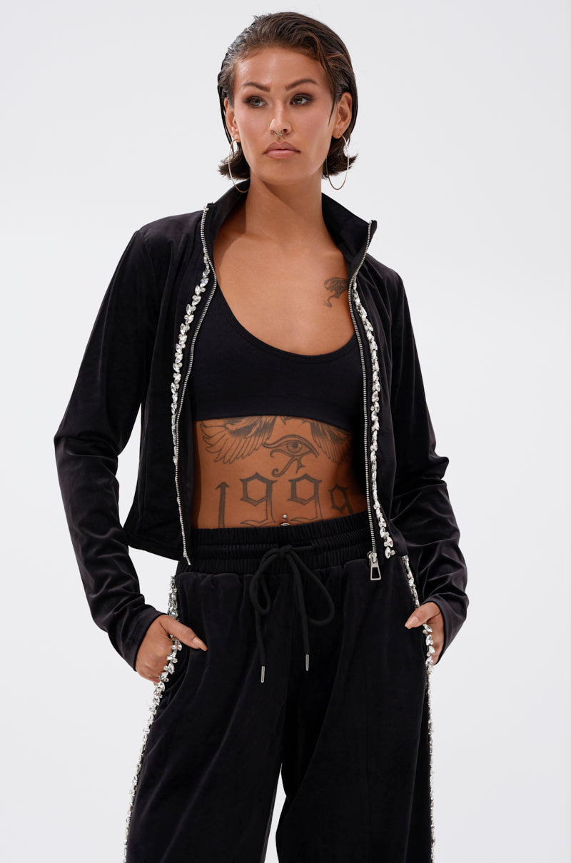 A person with short, slicked-back hair models the DARA RHINESTONE MOCK NECK ZIP-UP SWEATSHIRT over a matching crop top and pants, showcasing chest and stomach tattoos for an edgy vibe, hands casually in their pockets.