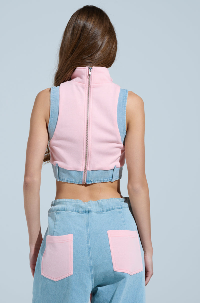 A person with long hair is seen from behind in the HEADS UP QUARTER ZIP SLEEVELESS SWEATSHIRT IN PINK, paired with denim pants featuring pink back pockets, all set against a light blue background.