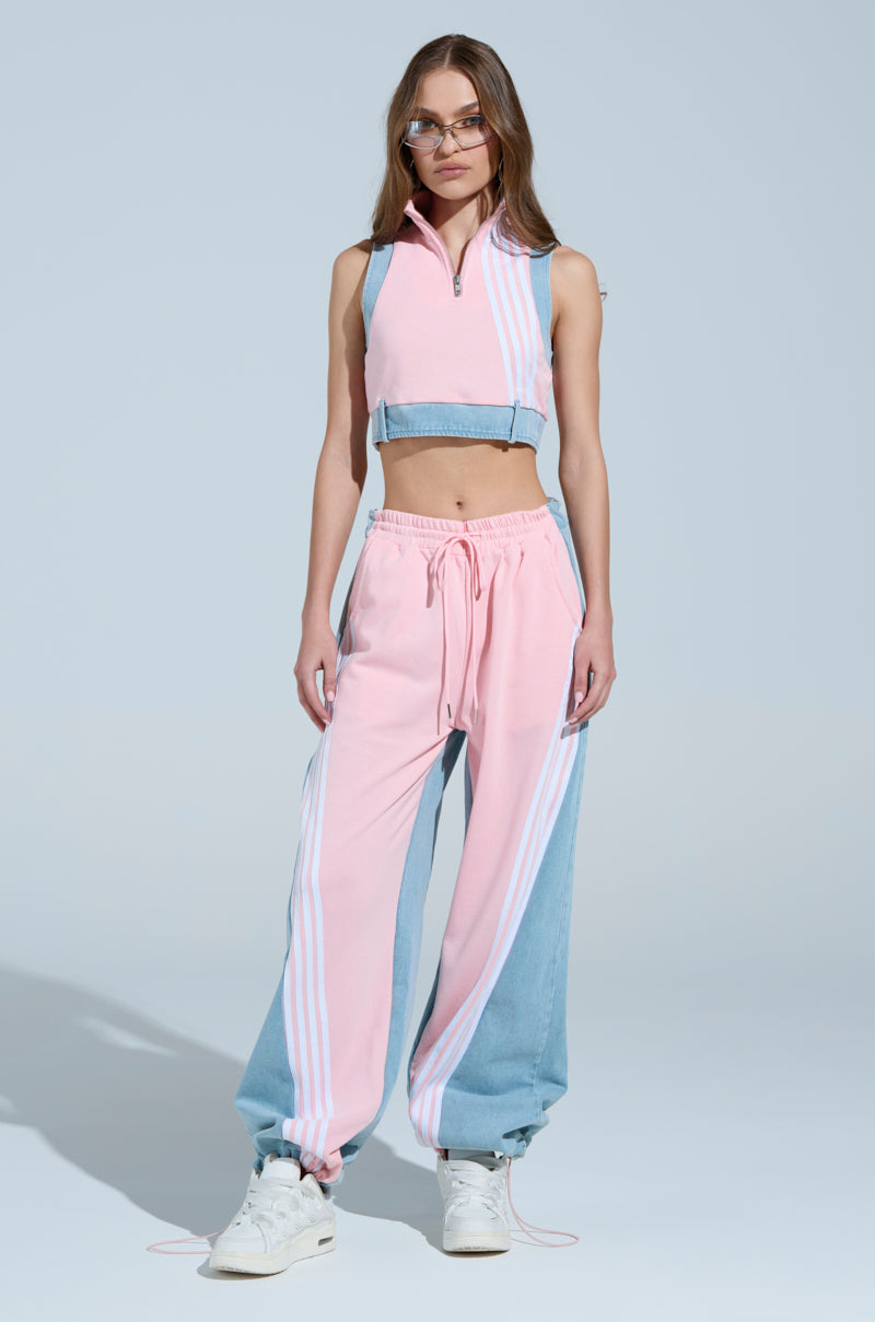 A woman poses in a stylish athleisure outfit against a light blue background, featuring the HEADS UP QUARTER ZIP SLEEVELESS SWEATSHIRT IN PINK and drawstring pants with athletic stripes, accessorized with sunglasses and white sneakers.