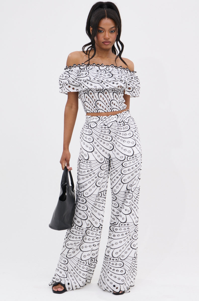 A woman poses against a white background in the SET YOU FREE OFF THE SHOULDER BLOUSE and wide-leg pants with a bold black and white abstract stitched pattern. She carries a black handbag and wears black open-toe heels.