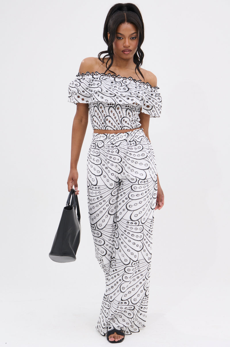 A woman stands against a white background, wearing the SET YOU FREE OFF THE SHOULDER BLOUSE and wide-leg pants with a black and white abstract stitched pattern, paired with black sandals and a matching handbag.