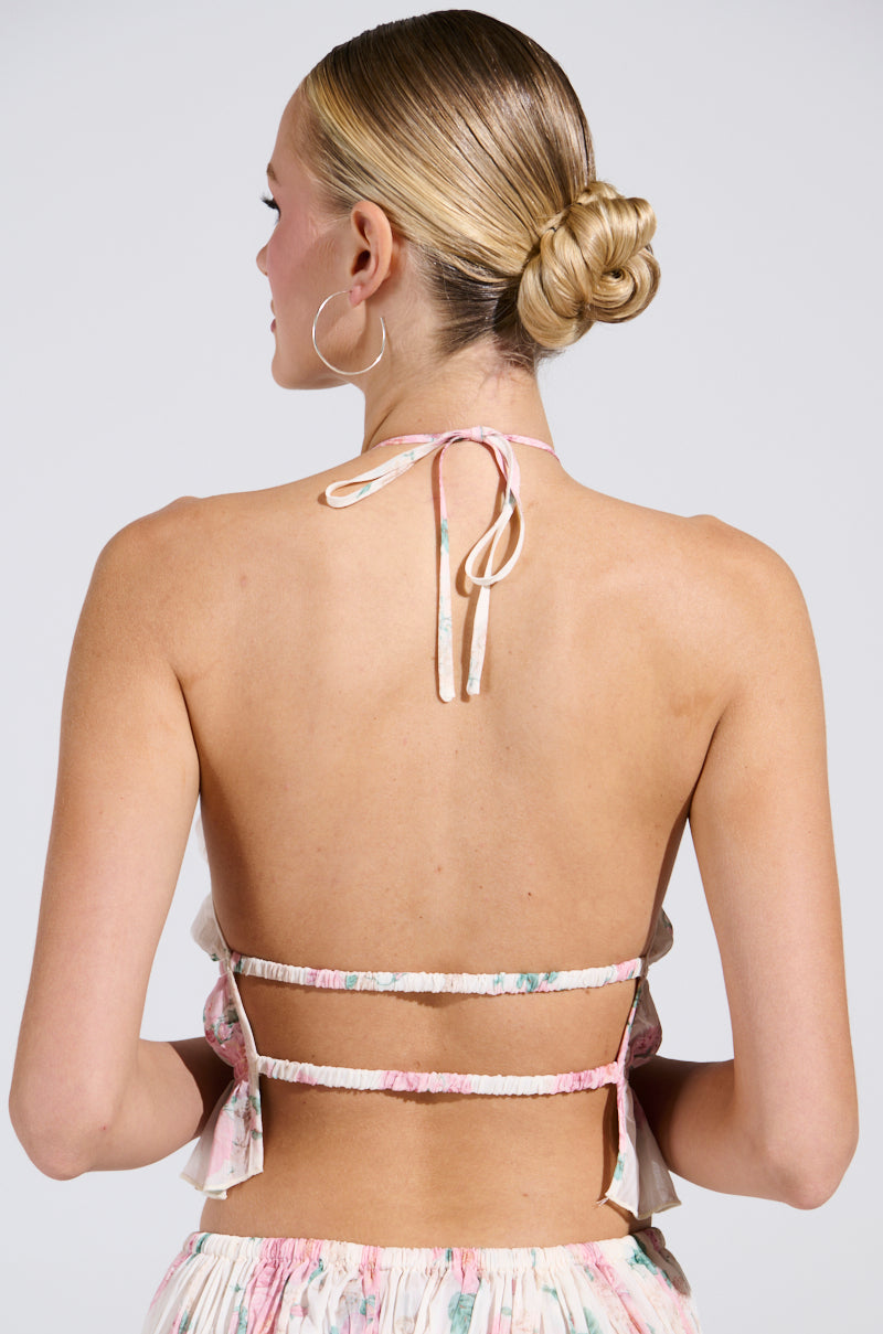 A person with a neatly styled bun models the RUBY FLORAL BLOUSE by AKIRA Label, featuring a floral pattern and two thin back straps. The cropped style exudes summery vibes against a neutral background, enhancing the outfit's allure.