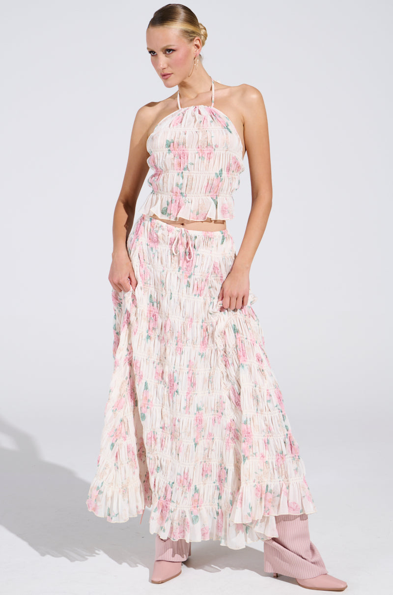 In a plain white studio, a person confidently poses in the AKIRA Label RUBY FLORAL BLOUSE, featuring a pastel floral pattern halter top paired with a matching flowy skirt, complemented by light pink ankle boots.