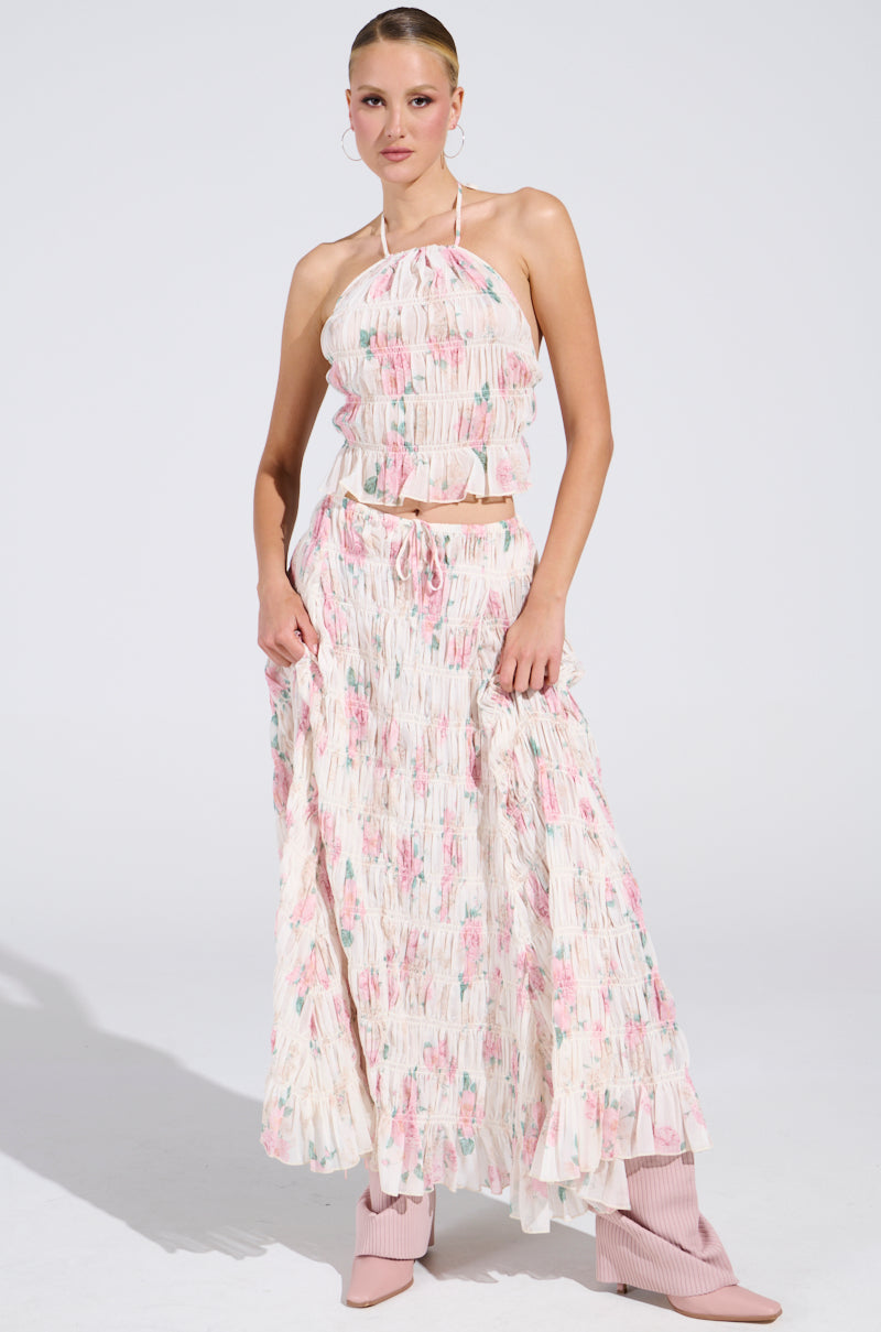 A woman confidently poses in the RUBY FLORAL BLOUSE with a cropped fit and soft pink and white tones, paired with a matching long skirt and light pink ankle boots.
