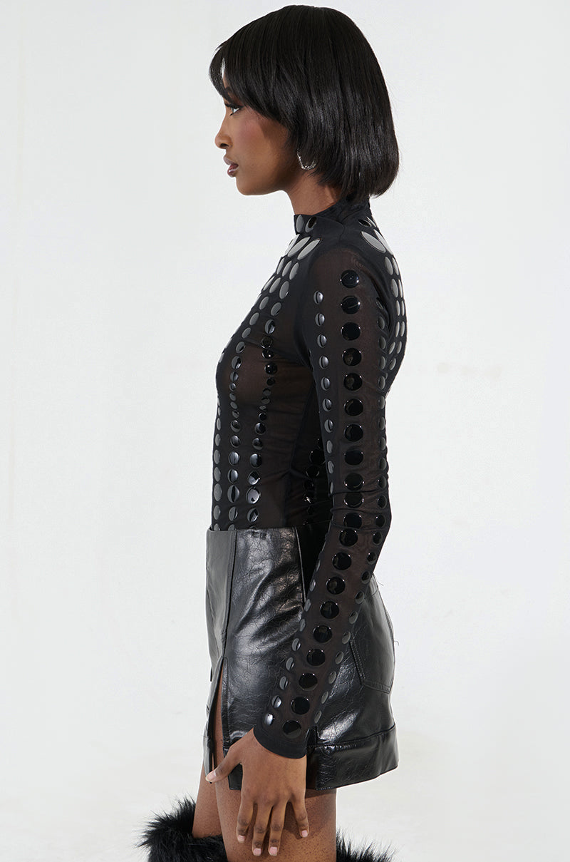 A person with short black hair stands sideways in a black outfit featuring circular patterns and a leather-like skirt. The NOT MY FAULT LONG SLEEVE MESH BODYSUIT adds depth to the reflective details, while the plain white backdrop highlights their striking silhouette.
