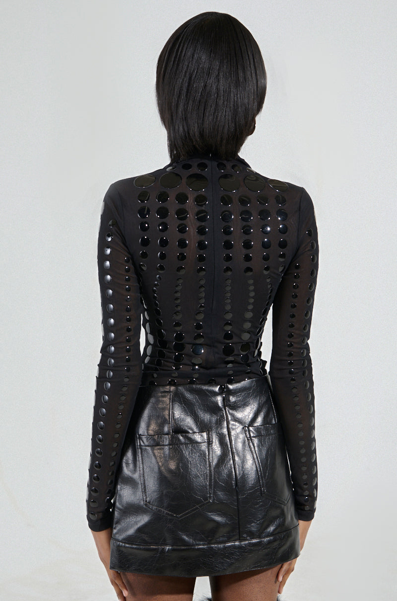 A person with medium-length hair is facing away, wearing a NOT MY FAULT LONG SLEEVE MESH BODYSUIT with textured circular patterns and reflective detailing, paired with a sleek black leather skirt. The background is plain and neutral.